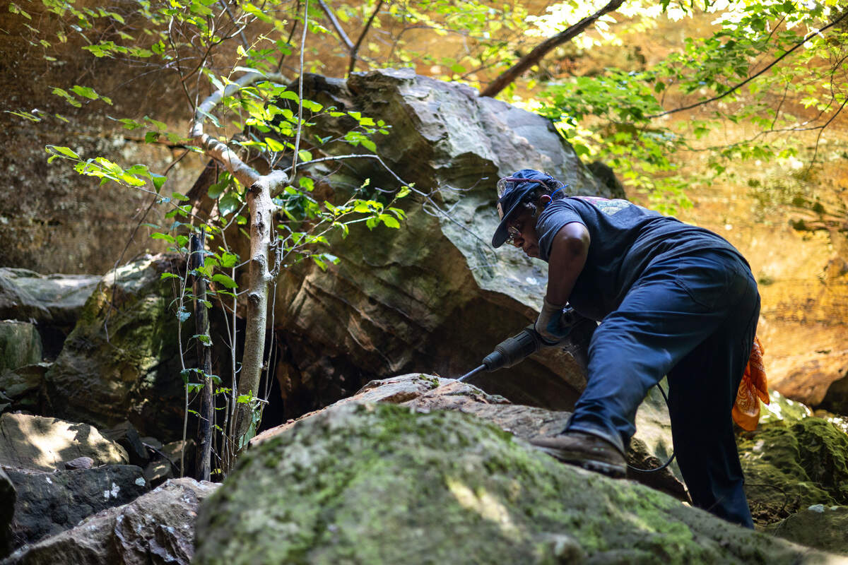 A woman in work boots, work pants, a t-shirt, a hat and glasses takes on a wide stance while standing on a rock. She uses a jackhammer to break apart the rock she is standing on. Greenery surrounds her.