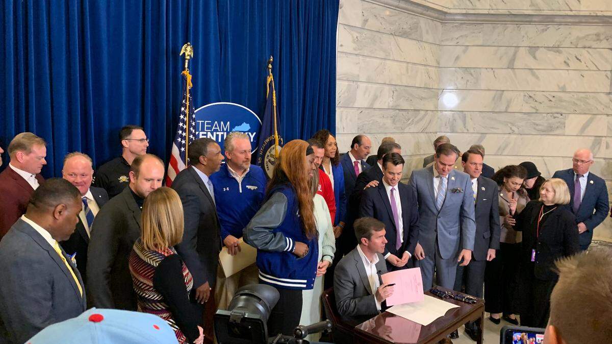 Gov. Andy Beshear is pictured signing Senate Bill 6 into law with several coaches, student athletes, administrators and lawmakers.