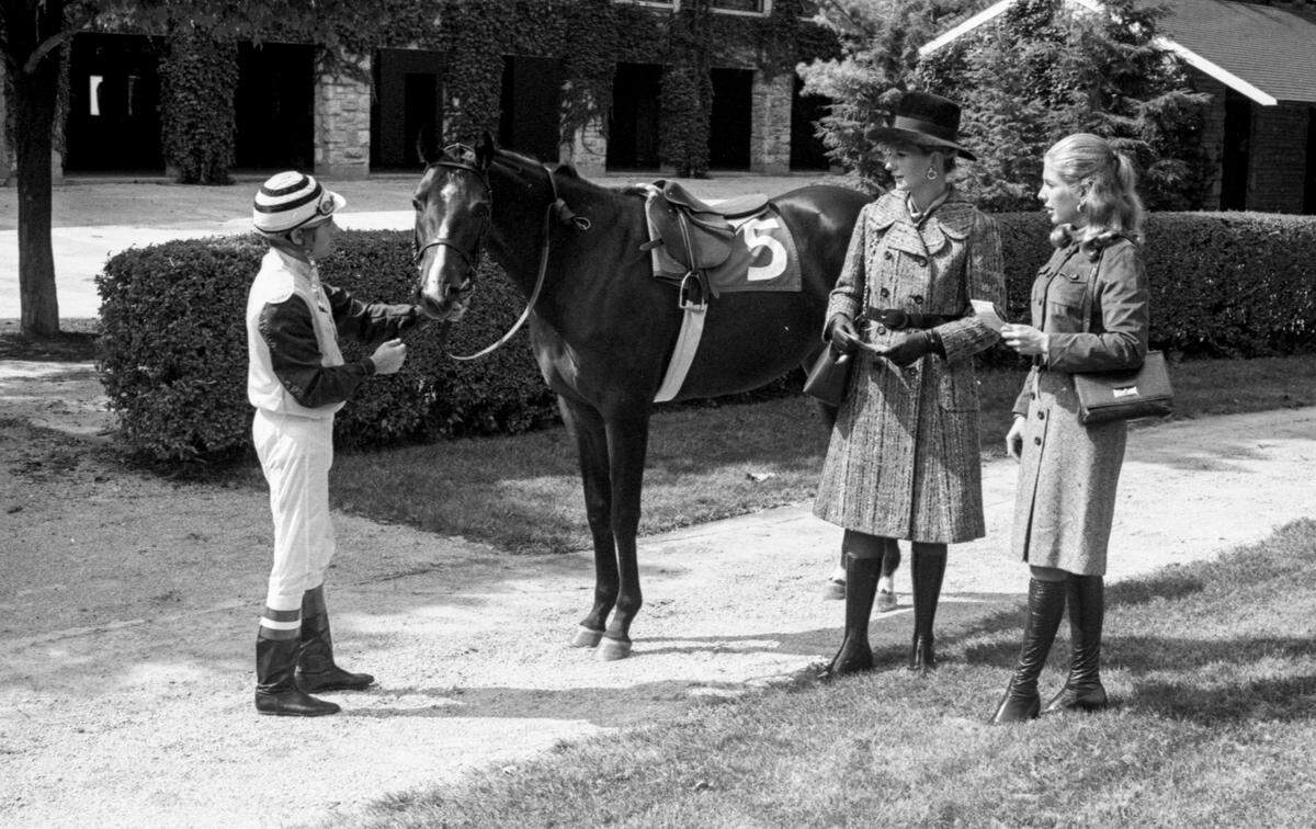 Fashion for the 1971 Keeneland Fall Meet at Keeneland Race Course in Lexington, Ky: Mrs. Hal Price Headley Jr., left, wears a black and brown slate combination semi-fitted tweed coat with notched lapels and brown suede belts by John Clark of Dublin from Wolf-Wile’s. Her hat, with wide brim, is of dark brown Italian velour by Mr. D. Her brown calf bag, with gold chain handle is by Korea. For a day at the races, Miss Mason Winn selected a dress with brown tweed skirt and gold jersey top with flattering roll collar. The costume is completed with a brown suede jacket with pocket detail by Helen Matthew. The boots are by Rosetta, all from Hymson’s. With the ladies is jockey Larry Rowe holding Lady Slipper from Lazy F. Ranch. Published in the October. 10, 1971 Sunday Herald-Leader.