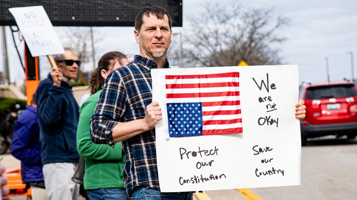 A protester holds a “We are not Okay” sign with the national flag upside down signaling America in distress before U.S. Rep. Andy Barr, R-Lexington speech on March 19, 2025, during the Commerce Lexington policy luncheon at the Hyatt Regency in Lexington, Ky.