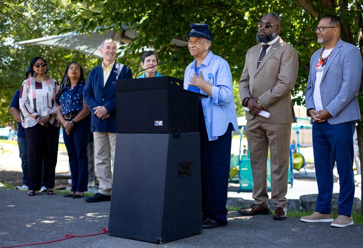 A woman wears a blue Civil War hat with gold lettering on the top while standing before a podium. A line of people reside behind her and listen.