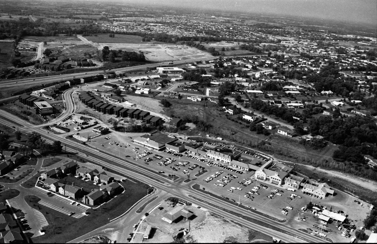 Aerial view of the Lansdowne Shopping Center in August of 1973, three years after it opened. Tates Creek Road runs diagonally near the bottom of the photo, while New Circle Road is seen cutting left to right in the top third of the image. A portion of the Merrick Place Apartments is at the lower left. Some of the businesses occupying the shopping center, from right to left, included, Lansdowne Exxon, The Lansdowne Shoppe liquor store, Norgetown Laundry & Cleaning, Big B Cleaners, Henri’s Fashions, First Federal Savings & Loan, Governor’s Table restaurant, Begley Drug Company, First Security National Bank and Trust Company, Hallmark Colony of Cards, Pet Gallery, Lexington Academy of Dance, Stretch & Sew Fabrics, Angelucci’s men’s ware, Gingiss Formalwear, Lansdowne Florist, Line and Shot sporting goods, Lansdowne Veterinary Clinic, Lexington Travel Center, Lansdowne Barber Shop, Timothy beauty salon, Baskin-Robbins Ice Cream, and the A&P grocery store.