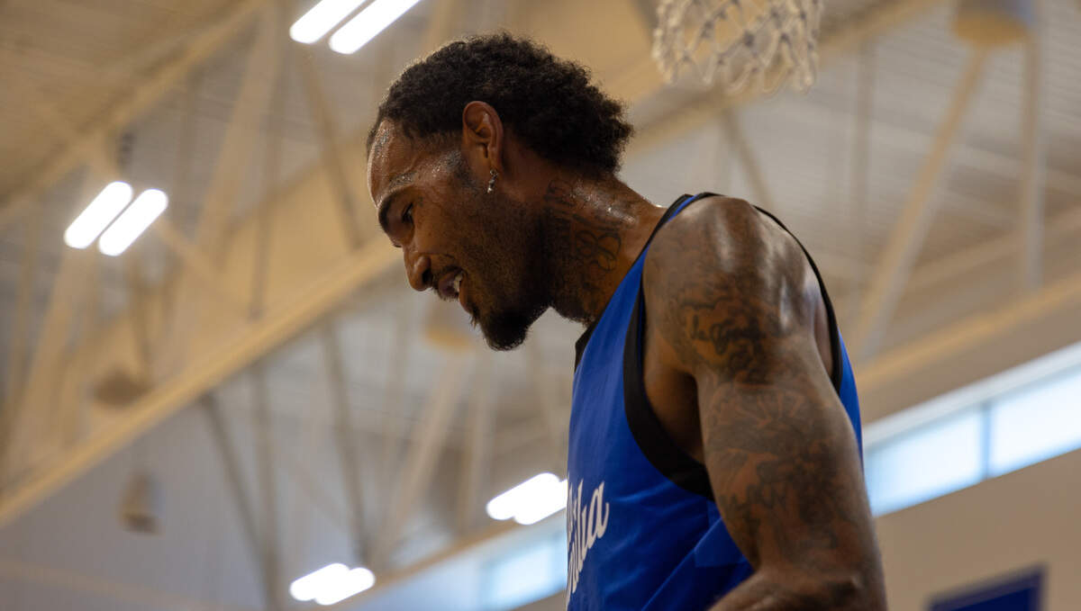 A basketball player with tattoos on his face, neck and arms wears a blue jersey and looks to the ground. Sweat has formed on his face.