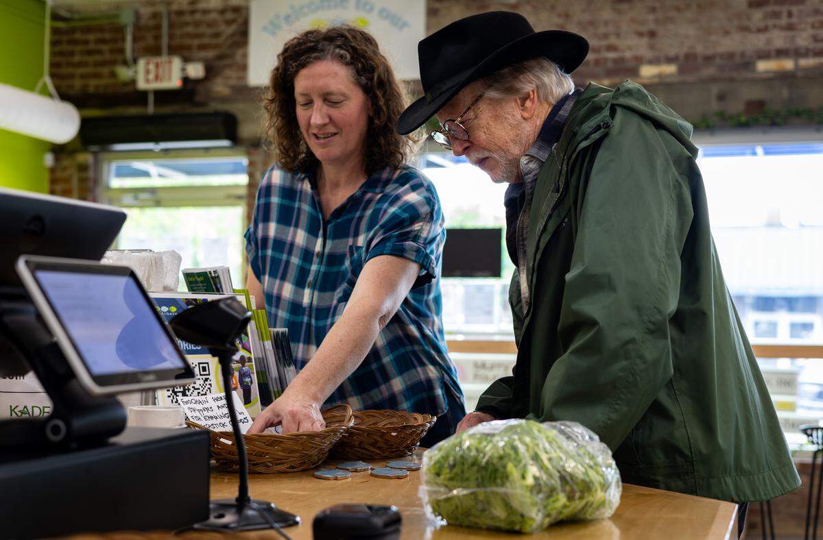 An elderly man, wearing a green rain jacket and a black cowboy hat, checks out his groceries, including lettuce, from the FoodChain Neighborhood Green Grocery while a woman speaks to him about locally made items for sale near the register. She is pointing to a weaved bowl containing the items while the man looks to where she is pointing.
