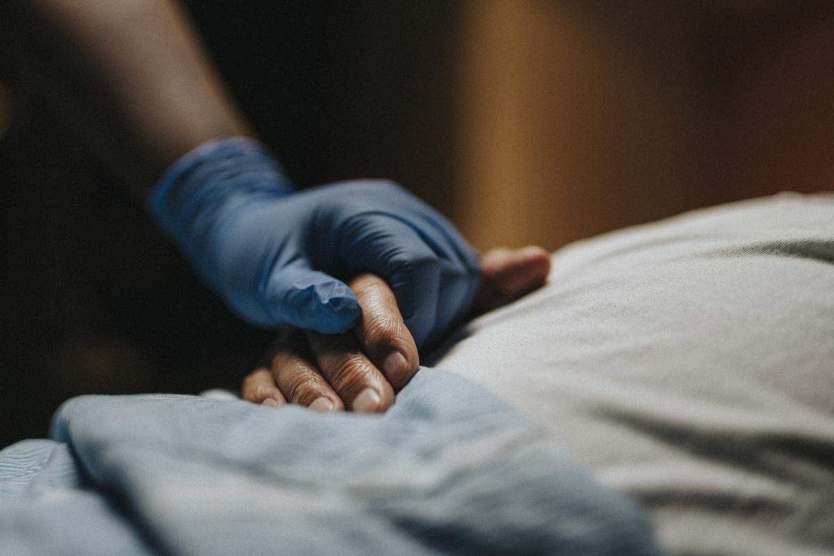 A gloved hand grasps a patient’s hand in a hospital setting.