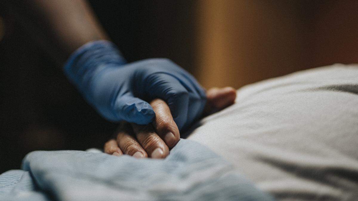 A gloved hand grasps a patient’s hand in a hospital setting.