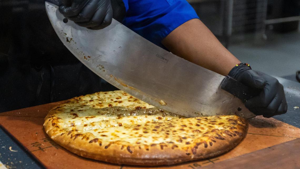 A cheese pizza from The Local Slice restaurant being cut into slices within Champions Kitchen dining area at University of Kentucky’s Student Center, October 26, 2023.