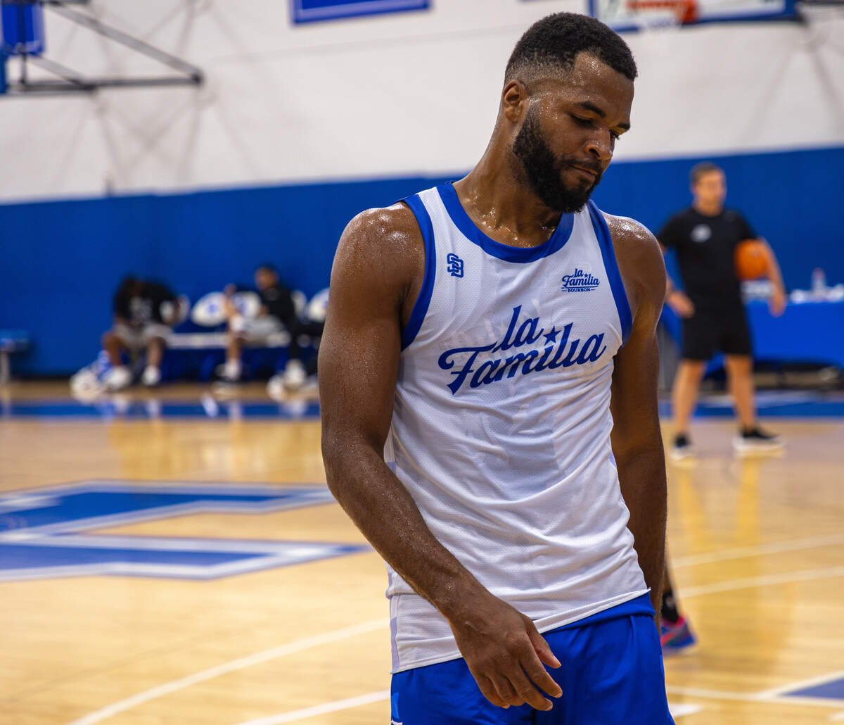 A basketball player wearing a white jersey and blue shorts stands on a basketball court, drenched on sweat.