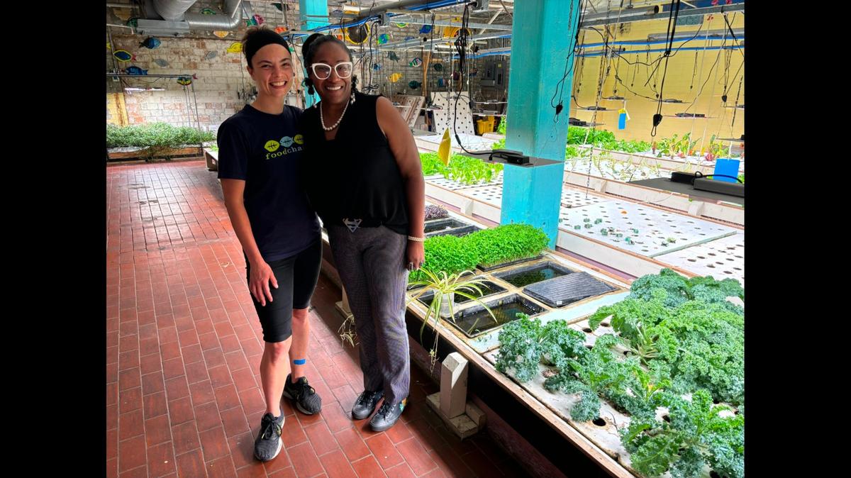 Leandra Forman (left) and Ka’Sondra Brown with FoodChain pose next to the nonprofit’s hydro and aquaponics farm. Beds of leafy greens can be seen under grow lights behind them.