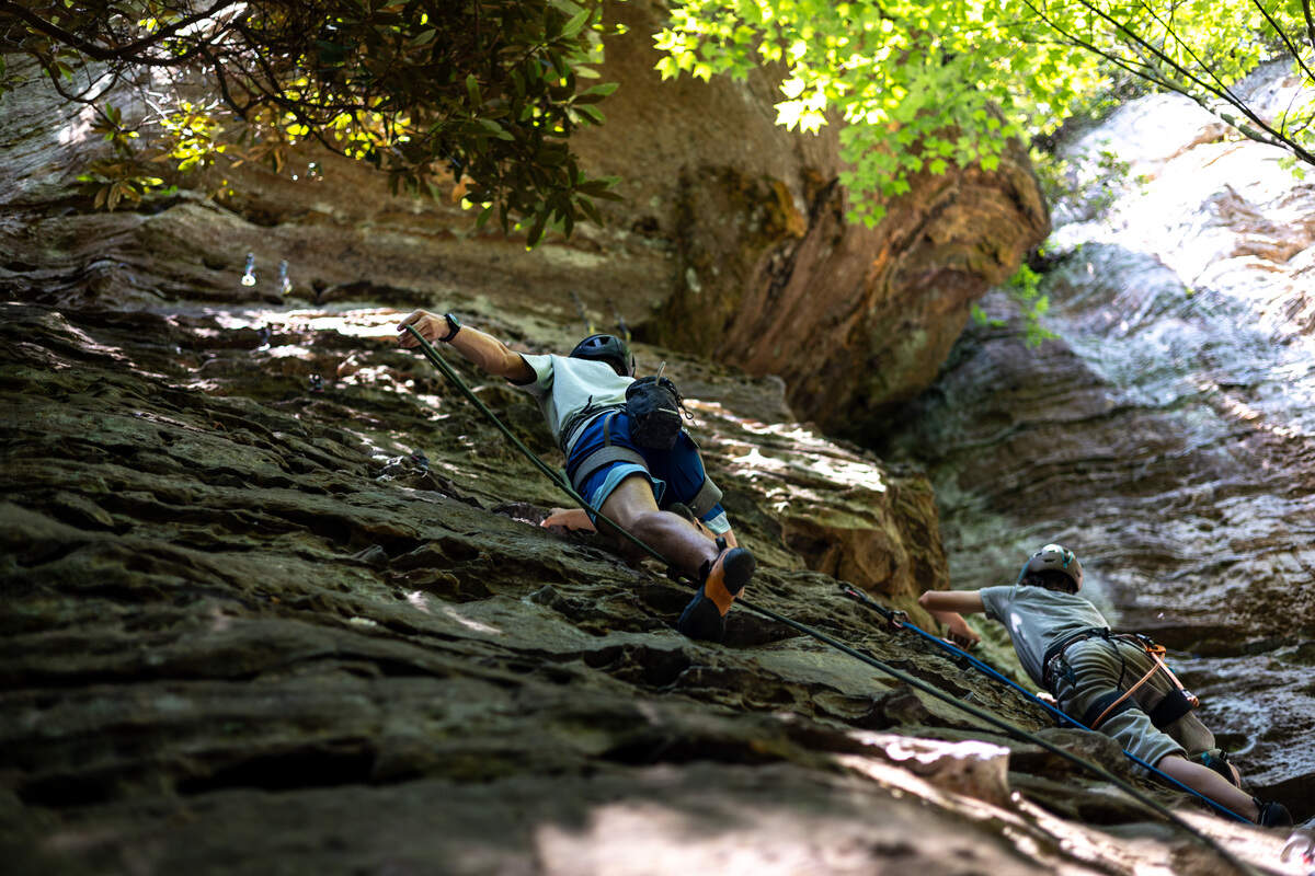 Two boys, both in gray shirts, climb a rock wall while attached to ropes. The trees around provide shade to the climbers.