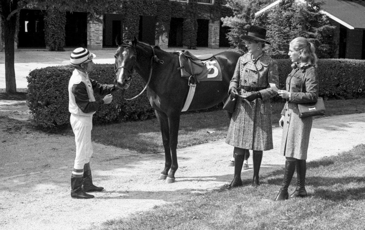 Mrs. Hal Price Headley Jr., left, wears a black and brown slate combination semi-fitted tweed coat with notched lapels and brown suede belts by John Clark of Dublin from Wolf Wile’s. Her hat, with wide brim, is of dark brown Italian velour by Mr. D. Her brown calf bag, with gold chain handle is by Korea. For a day at the races, Miss Mason Winn selected a dress with brown tweed skirt and gold jersey top with flattering roll collar. The costume is completed with a brown suede jacket with pocket detail by Helen Matthew. The boots are by Rosetta, all from Hymson’s. With the ladies is jockey Larry Rowe holding Lady Slipper from Lazy F. Ranch. 