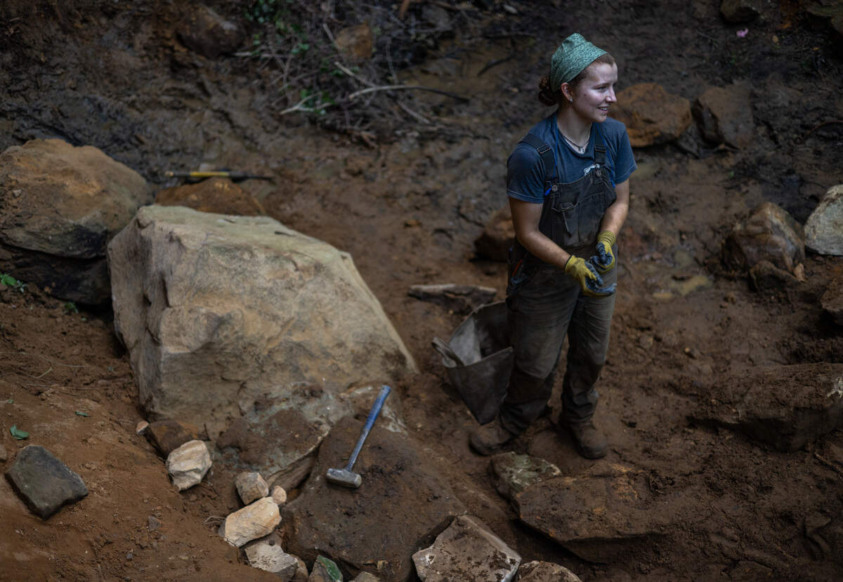 A woman wearing overalls, a blue t-shirt and a bandana on her head stands on mud and dirt next to a pile of rocks in a line.