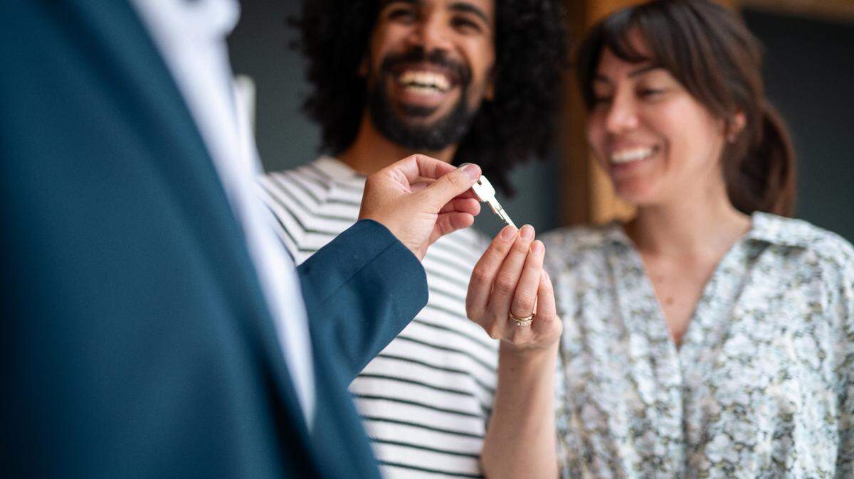 In this stock image, a real estate agent hands over keys to a smiling diverse couple, excitedly starting a new chapter in a modern home setting. They wear casual clothes, reflecting the relaxed nature of the milestone moment.