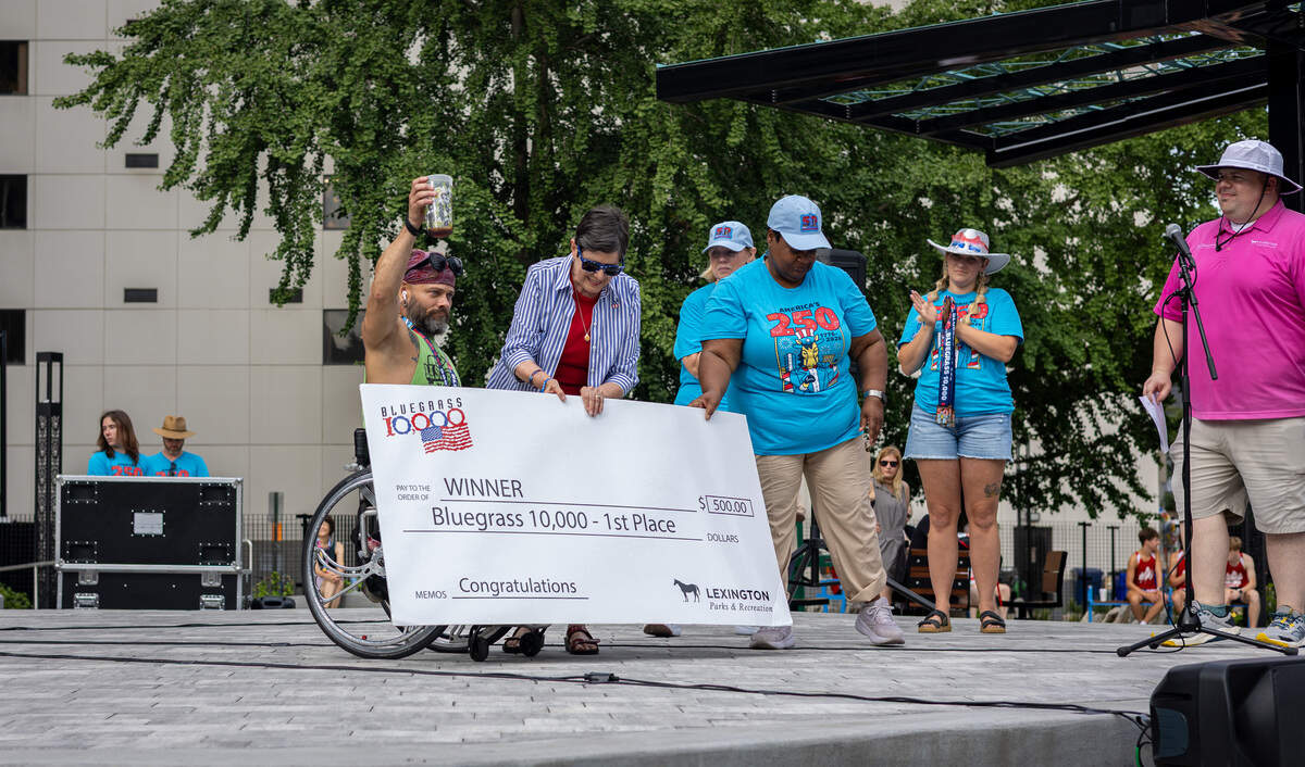 A man in a wheelchair and green tank top holds a beer in the air with his right hand. A lady next to him hands him a large check for $500.