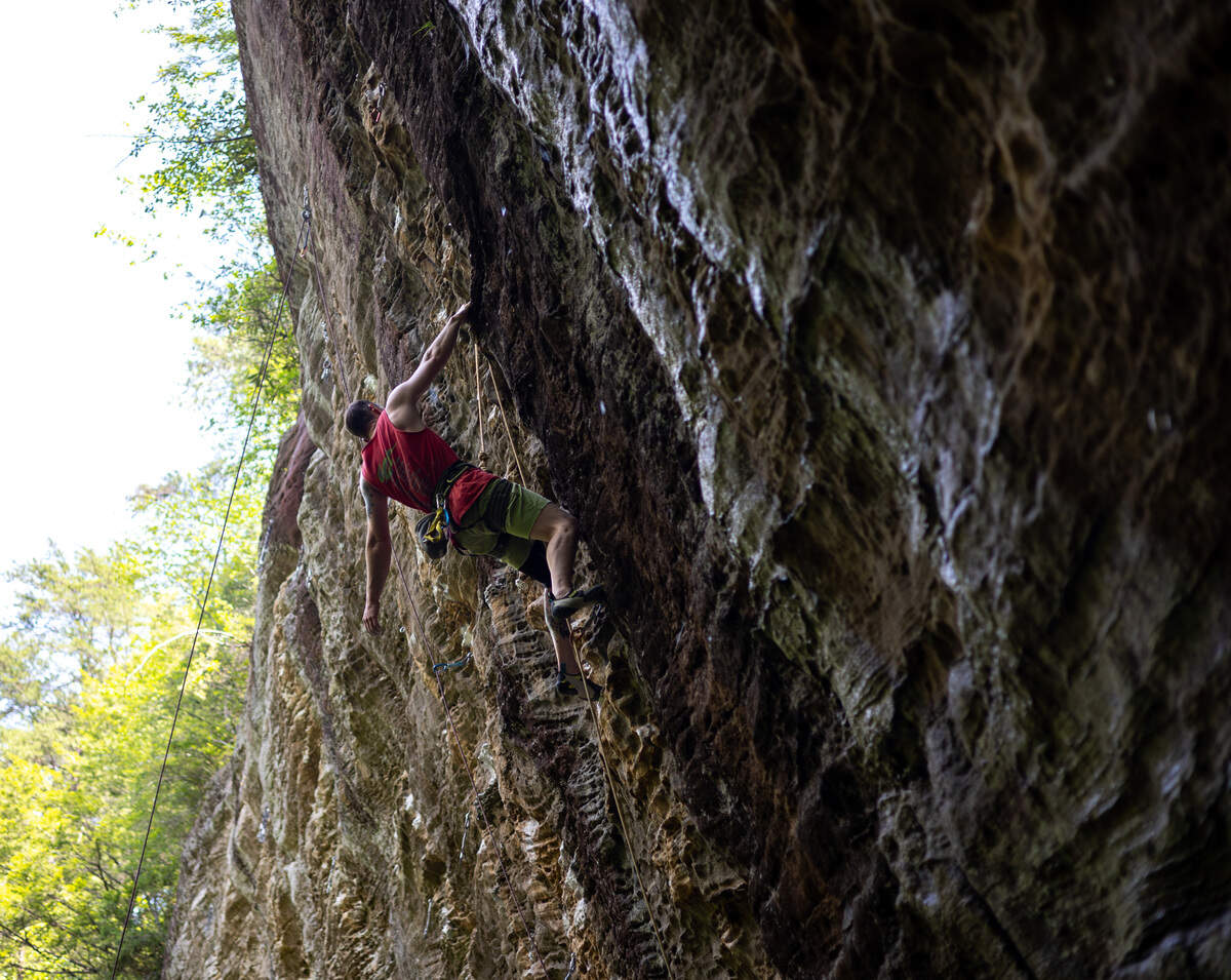 A man wearing a red shirt rock climbs on a vertical wall with one arm outstretched. He is attached to a yellow rope.