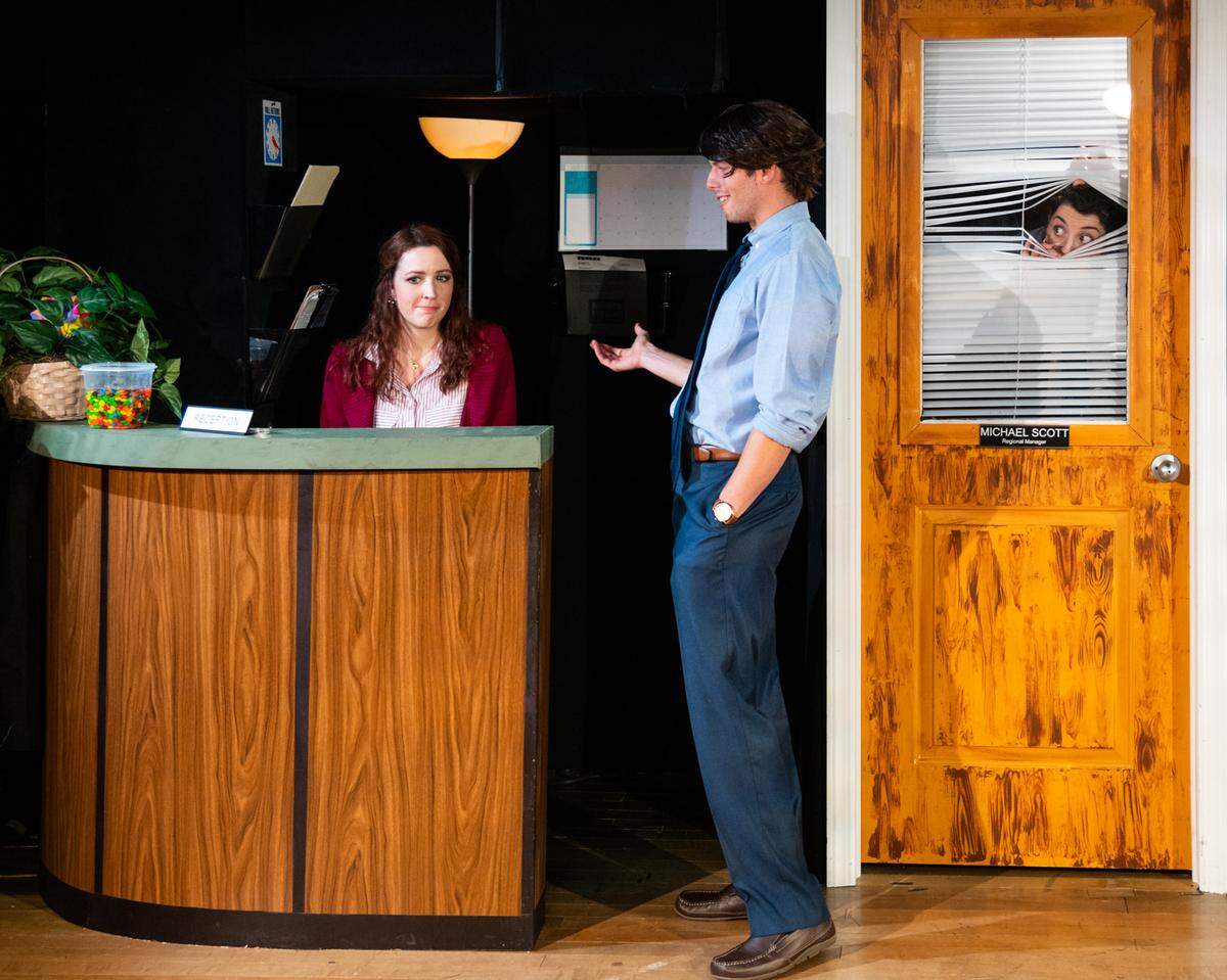 Madeline Glenn Thomas, as Pam Beasley, talks with Patrick Constant as Jim Halpert as Emma Brock playing boss Michael Scott sneaks a peek during “The Office! A Musical Parody”.