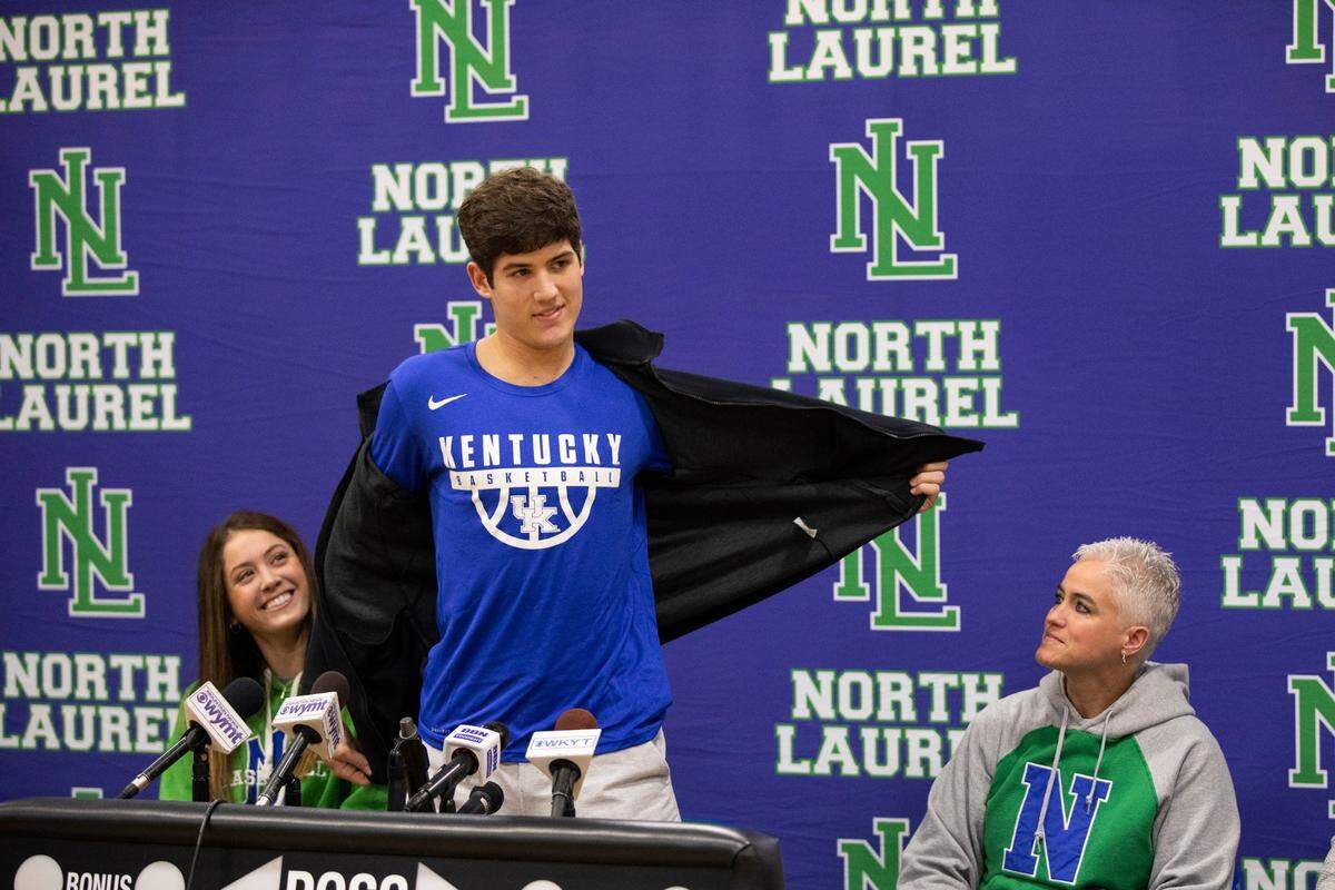 North Laurel High School basketball star Reed Sheppard takes off his jacket to reveal a University of Kentucky T-shirt upon announcing his college choice Saturday in London. At left is Reed’s sister, Madison. At right is his mother, Stacey, a former star player at UK.