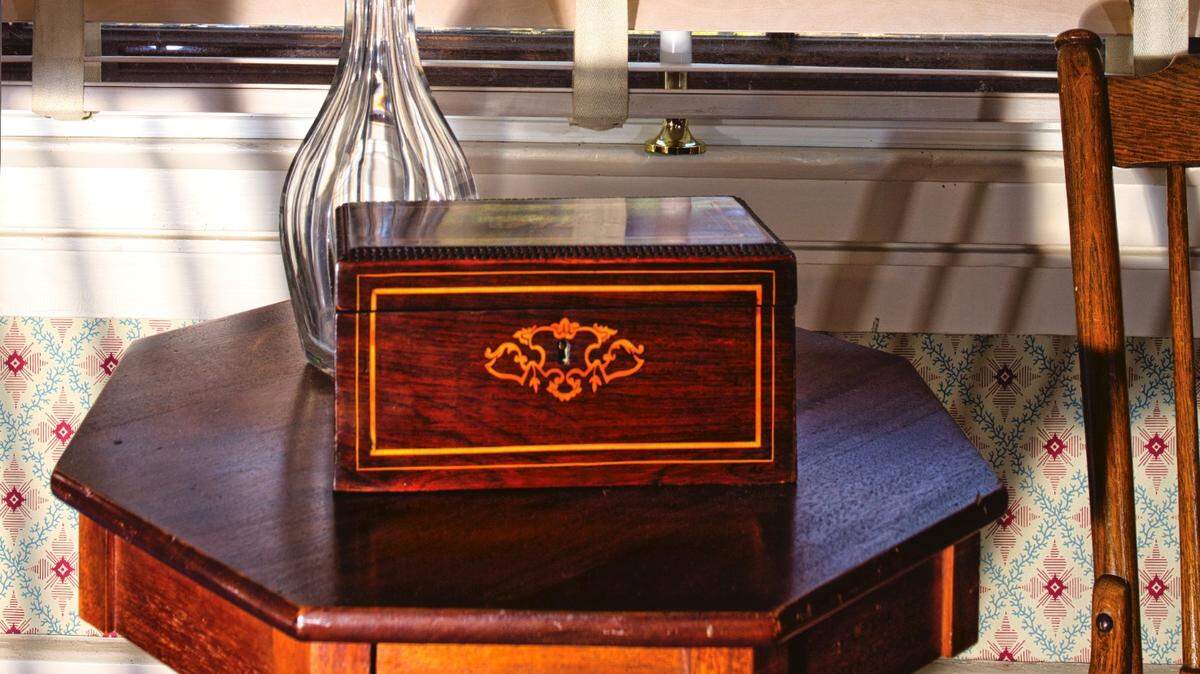 A wine decanter and tea caddy on octagonal rosewood table in the dining room, with the slave quarters through window. Photographer Bob Willcutt framed the image to contrast the lives of those who lived at Waveland.