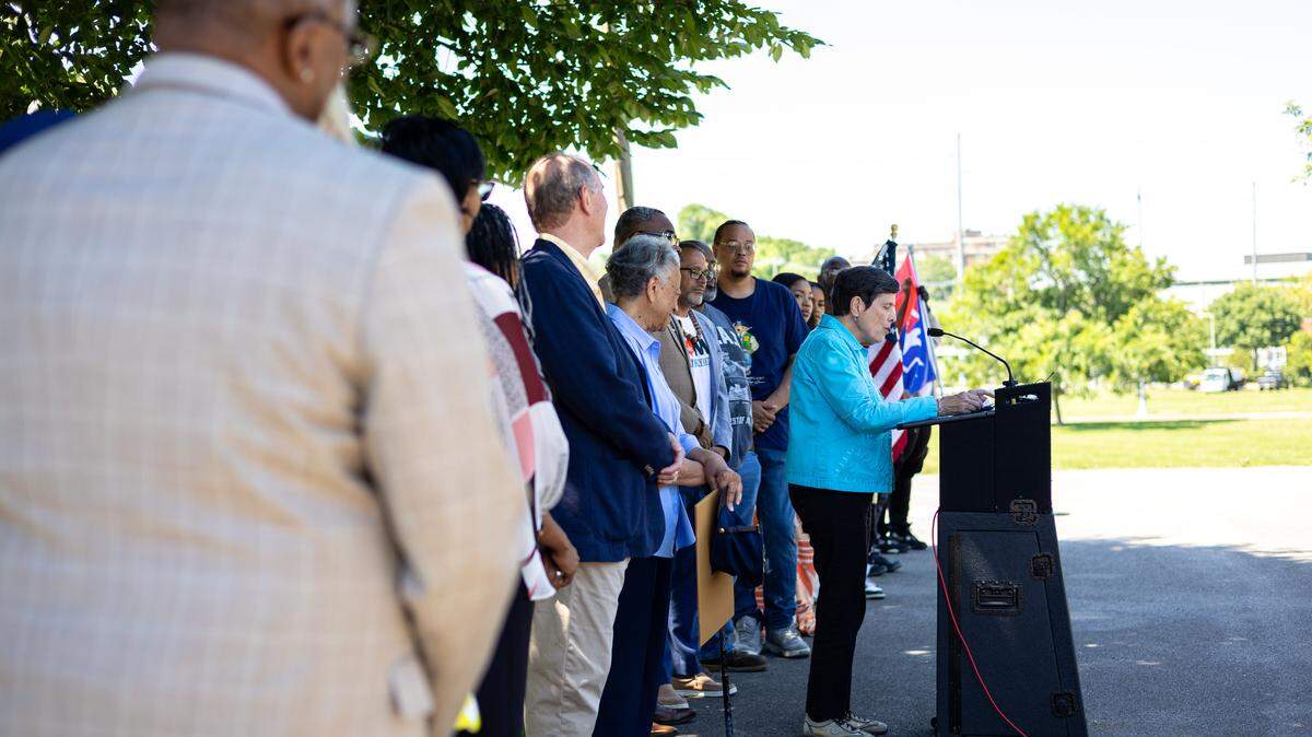 Linda Gorton, wearing a blue shirt, jeans, and white sneakers, stands in front of a line of people while talking into a microphone at a press conference.