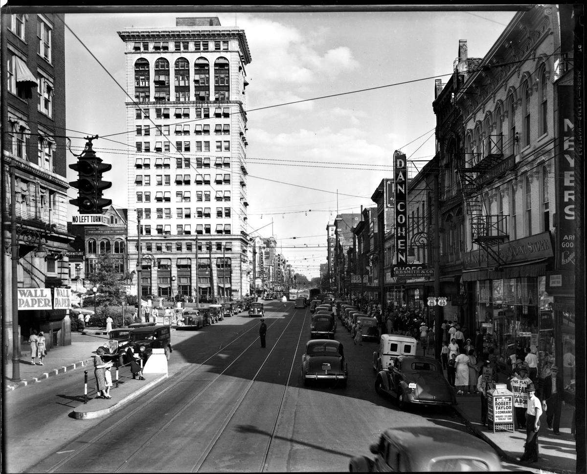Main Street, downtown Lexington, circa summer 1938. Rails run down the street, but around this time streetcar service was discontinued in Lexington. Like most American cities, as roads were improved and more people bought automobiles, trolley tracks were pulled up or paved over. Motor buses took over the steadily declining demand for public transportation. In the lower left corner of the picture you can see two women and a child waiting for the bus that coming towards them. Along the left side is what is now Henry A. Tandy Centennial Park. Further down is Lexington’s first skyscraper, the First National Building. Built in 1913, it is now a 21c Museum Hotel. The buildings on the right include a Woolworth five-and-dime store and clothing store B.B. Smith & Co., whose sign said it was “Correct Appareral for Women & Misses”. This is now the site of the Lexington Financial Center, or more commonly known as the “Big Blue Building”.