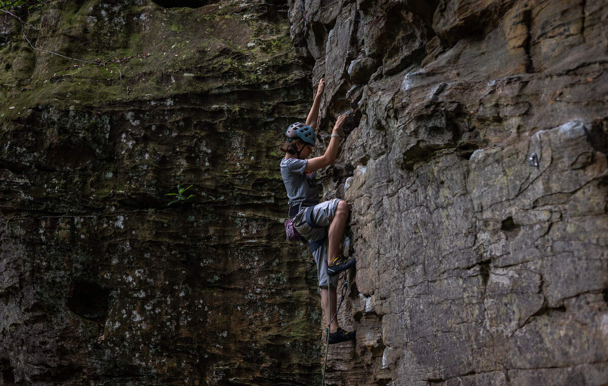 A person, attatche to a rope, wears a helmet and gray clothing while climbing a rock wall. The wall is a slate gray next to a mossy wall of rock.
