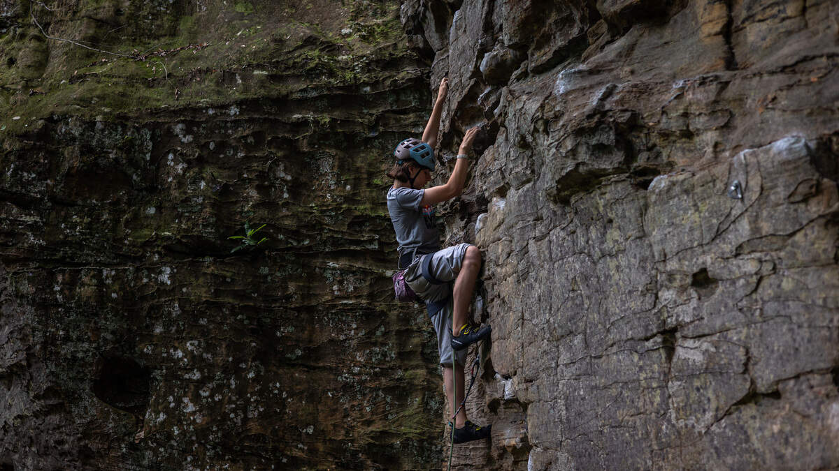 A person, attached to a rope, wears a helmet and gray clothing while climbing a rock wall. The wall is a slate gray next to a mossy wall of rock.