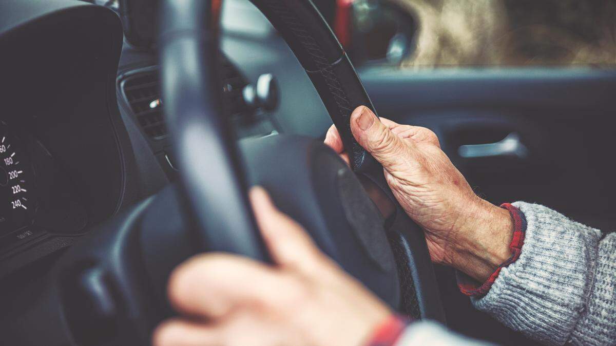 In this stock image, there's a close up of an elderly man's hands on a steering wheel. There's a depth of field effect where the man's right hand is in focus but everything else is blurred.