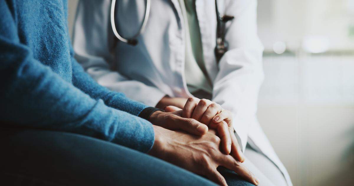 The stock image shows a doctor holding a patient’s hands in a medical setting.