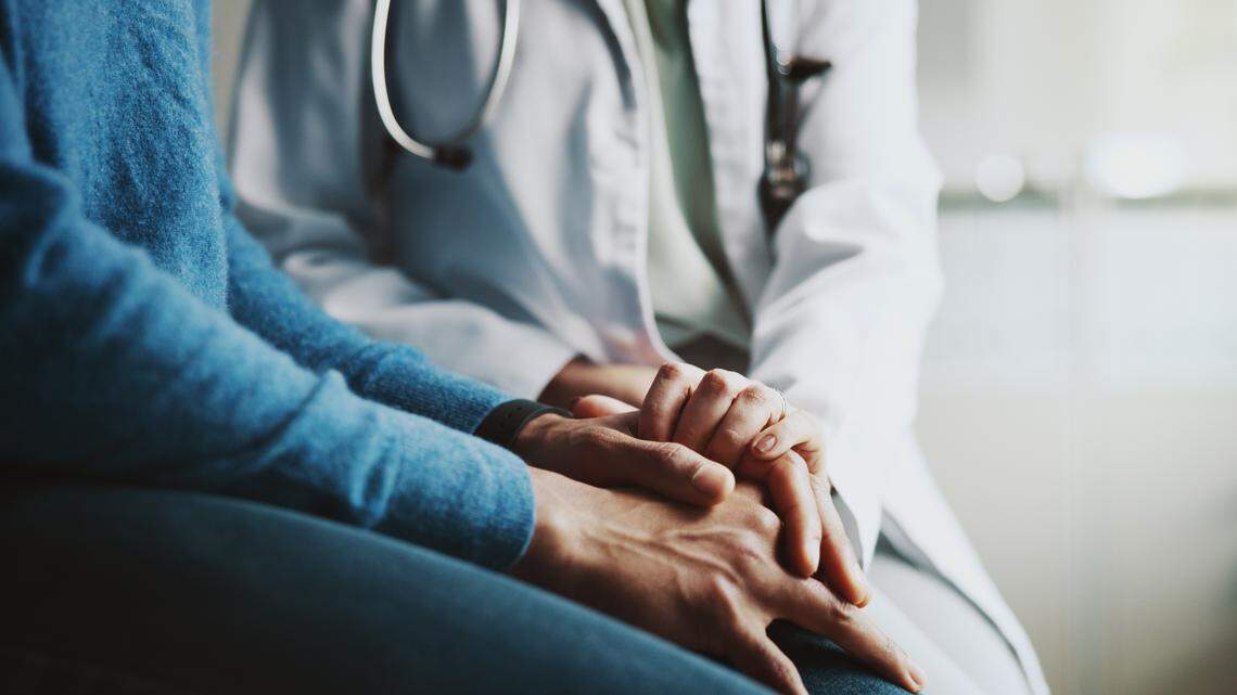 The stock image shows a doctor holding a patient’s hands in a medical setting.