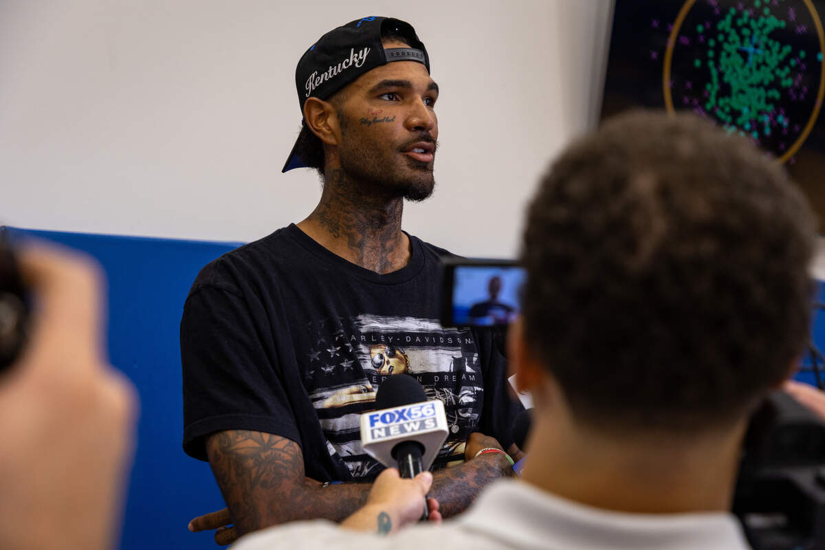 A basketball player with tattoos on his face, arms and neck wears a black t-shirt and backwards, black hat while talking to a group of 10 or more people holding microphones and cameras, all pointed at him. They are located on the sidelines of a basketball court where the bottom of the walls are covered in blue mats.