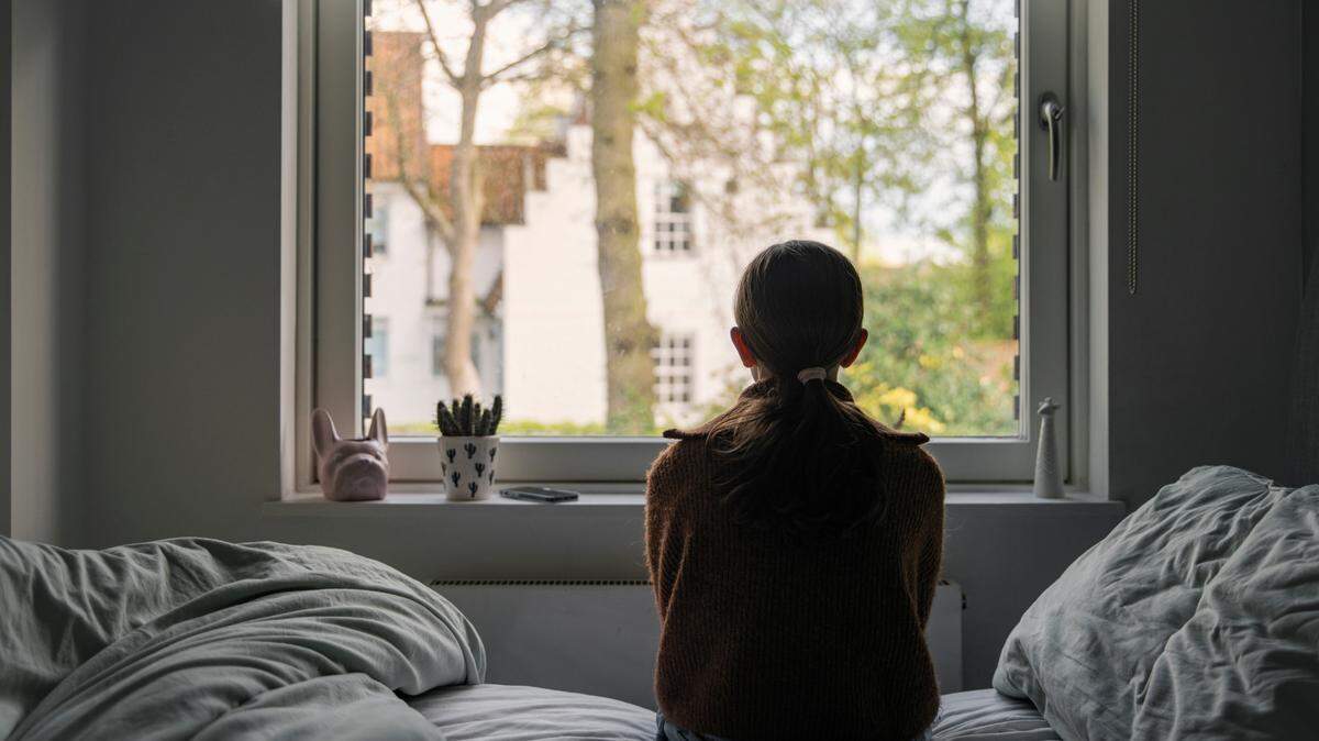 A young girl looks out of her bedroom window. The view through the window is blurry but shows a residential area.