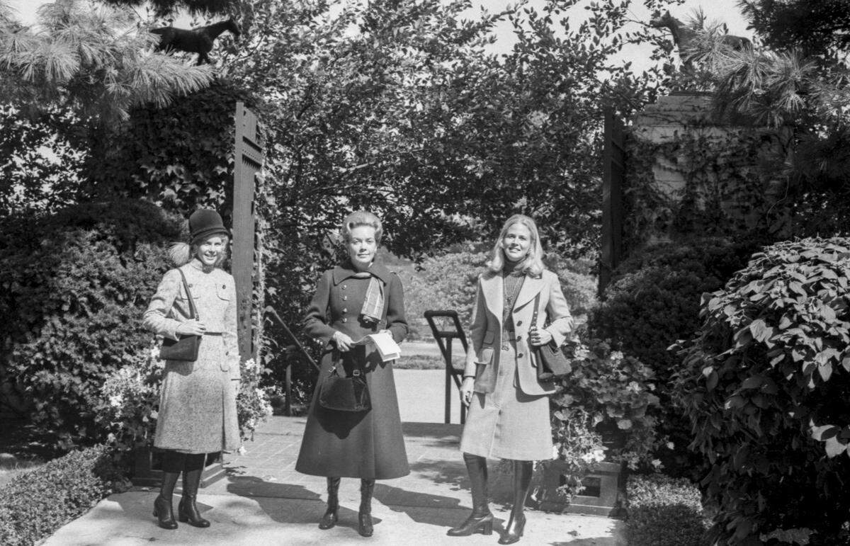 Mrs. John H. Hardwick, left, enters the track wearing a brown Donegal tweed dress and jacket by Dominic Pompollo. The dress features a brown jersey top with high collar from Ebro’s. Her hat is of brown velour with the Chanel profile. Mrs. Barrett H. Morris, center, selects a double-breasted midi coat in deep green with brass button detail by Peabody House from Four Season. Mrs. David Parrish is seen in a Jaeger skirt and single breasted wide-lapel blazer, both camel. With it she wears a gray cashmere turtleneck sweater by Pringle of Scotland from Meyers. 