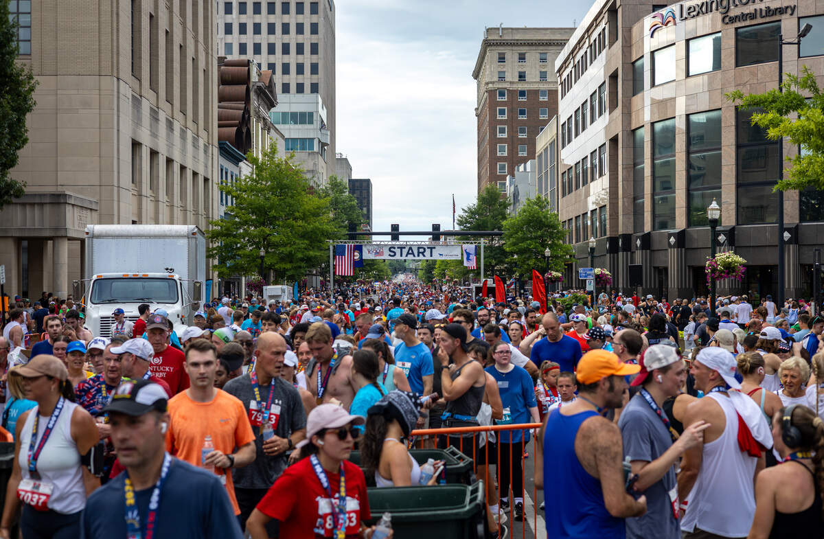 A large crowd of people stand on a street. A large sign that overhangs the street says “start” off in the distance. Large buildings are on the side of the street.