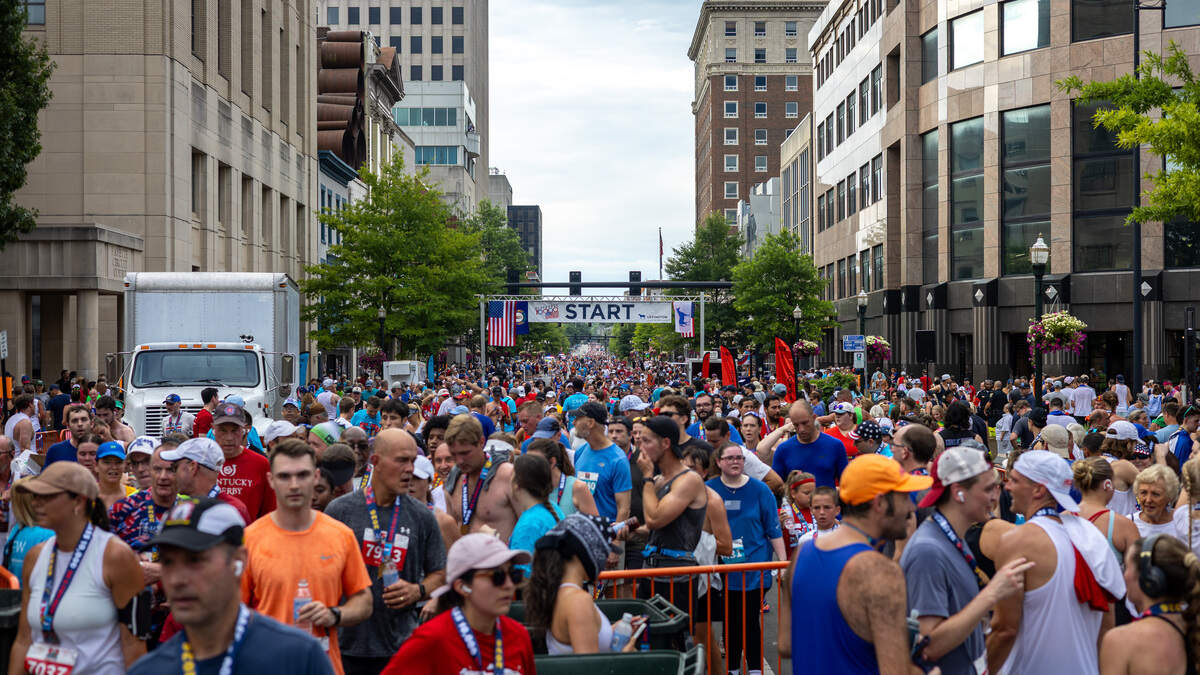 A large crowd of people stand on a street. A large sign that overhangs the street says “start” off in the distance. Large buildings are on the side of the street.