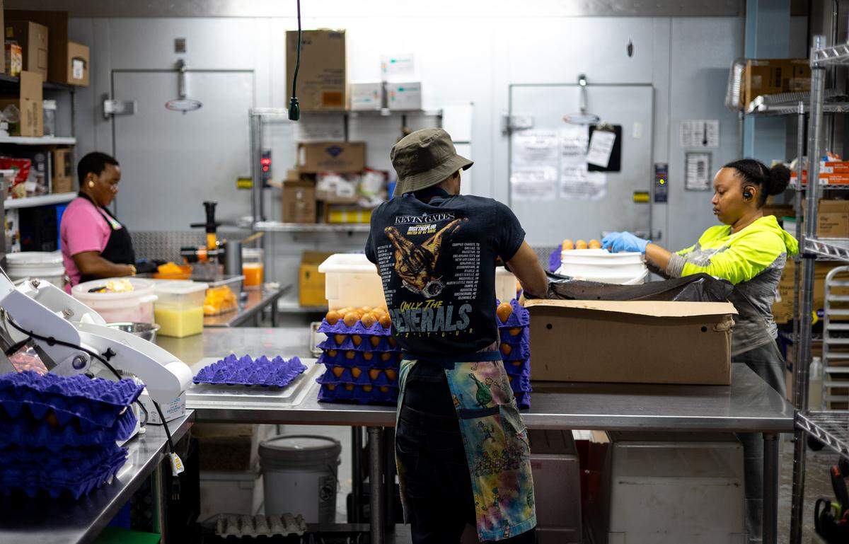 A man in a black t-shirt and tie-dyed apron, in the center of the frame, and a woman in a neon-yellow and gray jacket, on the right side of the frame, puts eggs into packaging. A woman on the left side of the frame in a pink shirt and black apron puts mangos into a blender to be made into mango juice. The kitchen is all gray, with gray walls and silver shelving. A cord dangles near the man in the center of the frame.