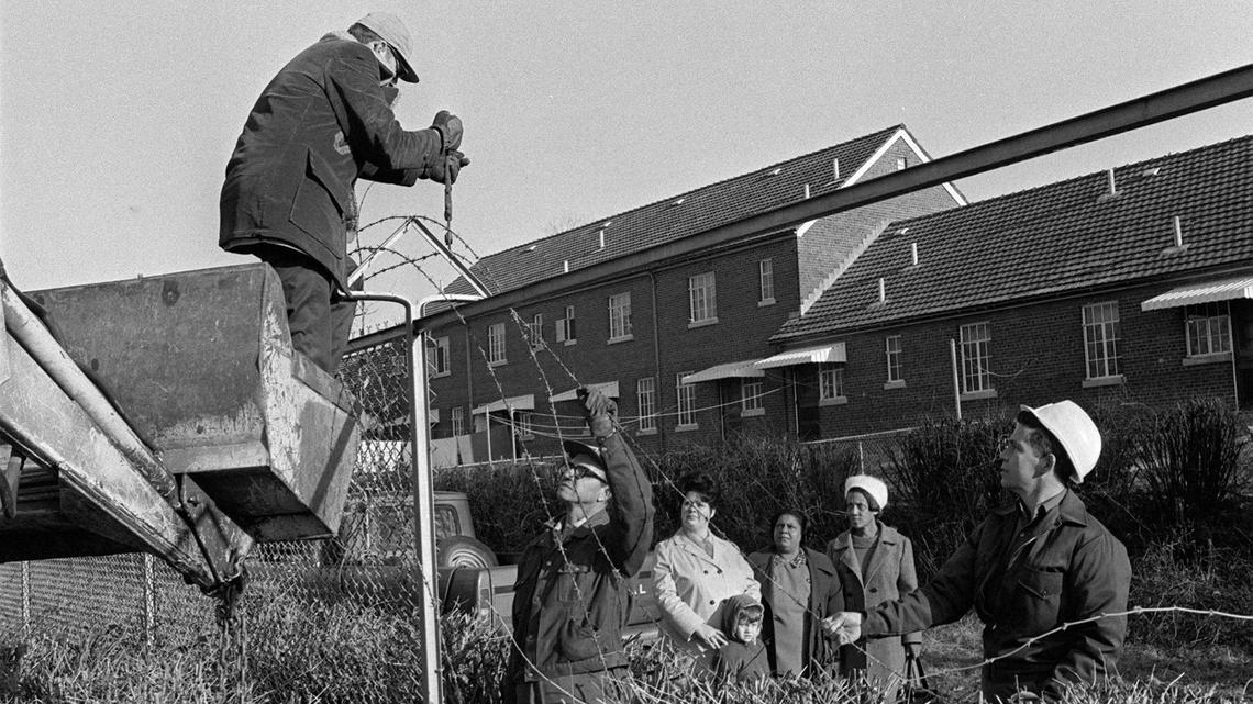 An eight-foot fence that separated blacks and whites since 1939 in the Blue Grass-Aspendale Housing Project comes down January 30, 1974, in Lexington, KY. Taking down the barbed wire are, left to right, Jay Martin, Warren Gerton and Earl Osten, supervisor. Looking on are, left to right, residents, Mrs. Ruby Anderson and her son Steve; Mrs. Edith Hines, president of the Residents’ Council and Mrs. Lola Jones, also a council member.