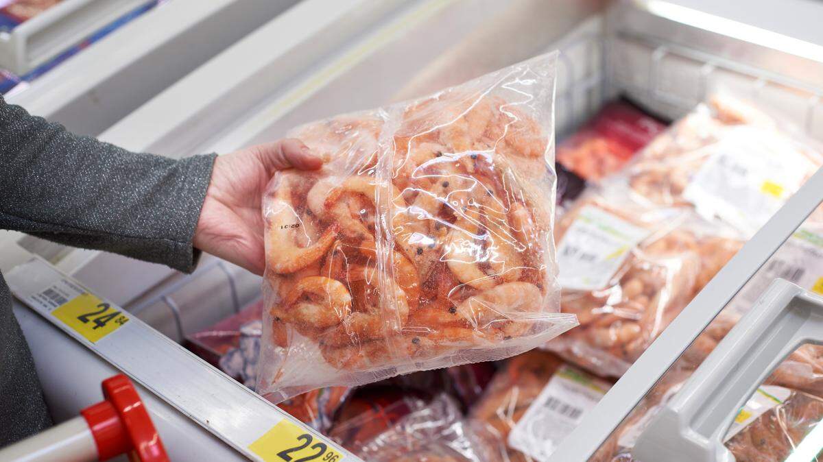 In this stock image, a person reaches into a grocery store’s freezer to pull out a bag of shrimp.