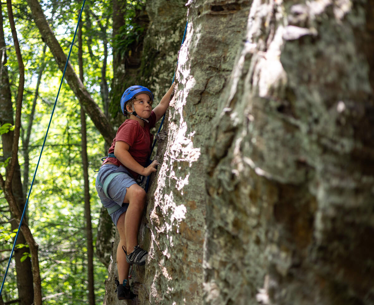 A young boy in a red shirt climbs a rock wall while attached to a rope.
