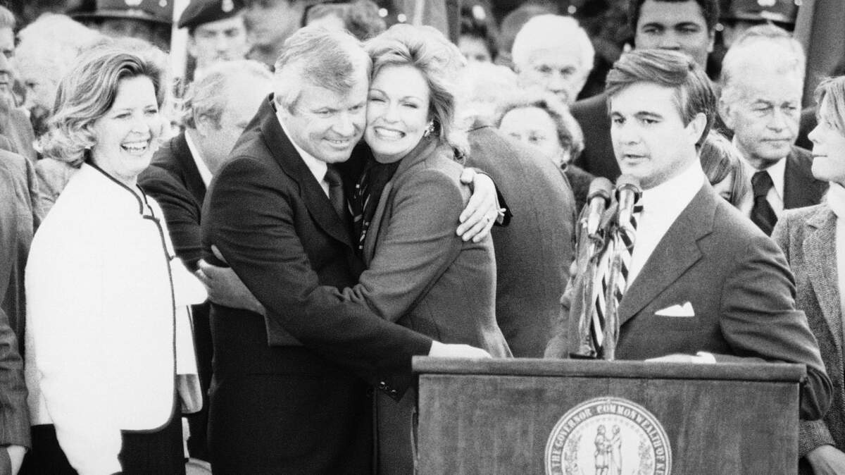 New Kentucky Governor John Y. Brown, Jr., hugs his wife, Phyllis George, after being sworn in as the 55th chief executive of the Commonwealth in Frankfort, Kentucky on Tuesday, Dec. 11, 1979. Looking on is the new Lt. Governor Martha Layne Collins, left. (AP Photo)