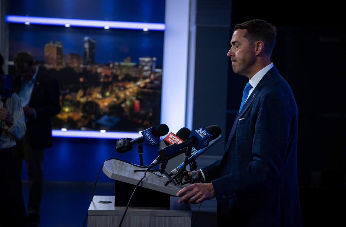 A man in a blue suit stands at a podium with white lights shining down on him. His eyes are closed, and his head is tilted down.